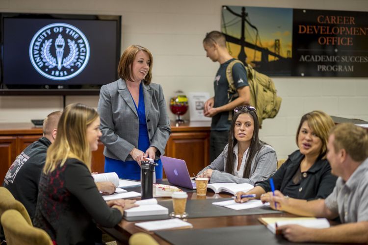 A woman in a gray suit and blue dress stands up while talking to a table of five seated people