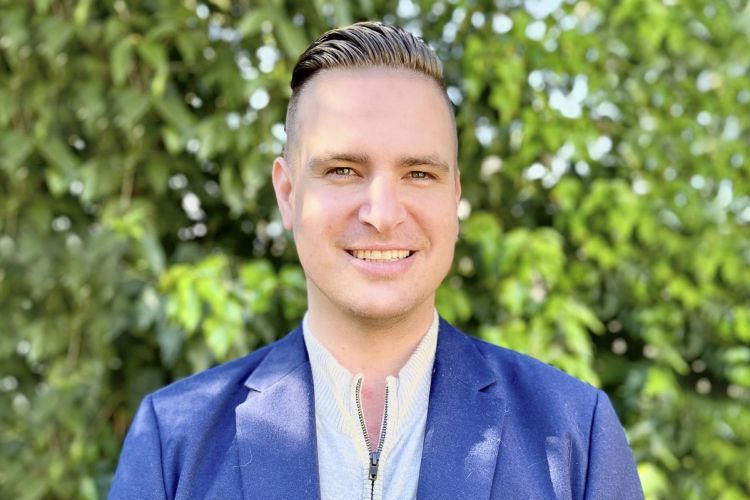 A man in a suit smiles in front of a wall of greenery