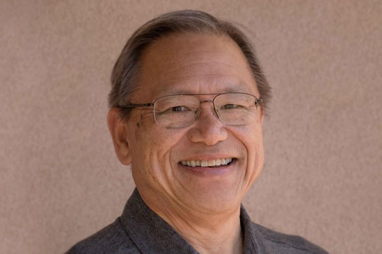A portrait of a man wearing a gray polo shirt in front of a tan neutral background 