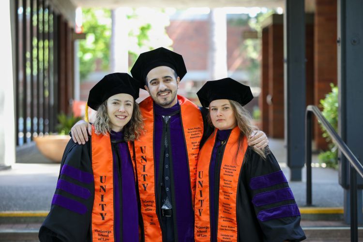 Four graduates wear purple and black robes with graduation caps on and orange sashes that say "UNITY McGeorge School of Law"