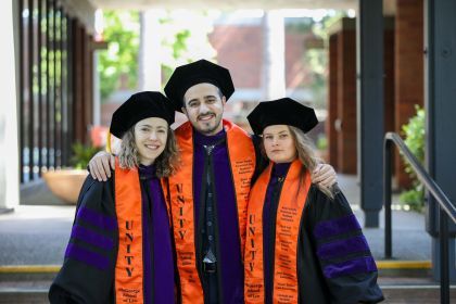 Four graduates wear purple and black robes with graduation caps on and orange sashes that say "UNITY McGeorge School of Law"