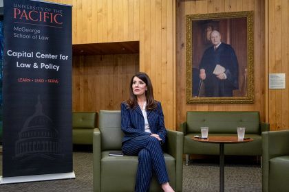 A woman in a dark blue stripped suit sits in a green chair in front of a sign that says "University of the Pacific McGeorge School of Law Capital Center for Law and Policy.".